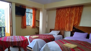 A hotel room with two beds, a small table, a TV mounted on the wall, and a balcony with orange curtains.