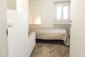 A small, single-bed room with a herringbone-patterned floor, a radiator, and a window with blinds.