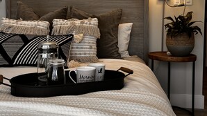 A neatly made bed with a tray of coffee cups and a French press, a wall-mounted lamp, and a framed artwork above the bed. - Upper Rideau - “The West” Resort (Portland)