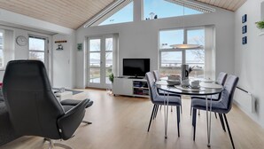 A modern living room with a large TV, a dining area with a round table, and large windows offering a view of the outdoors. - 3 bedroom amazing home in Mariager (Mariager)