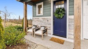 A house with a dark blue front door, a stone wall, and a wooden porch with two chairs.
