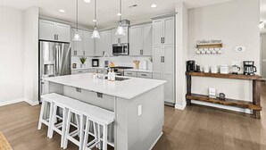 A modern kitchen with white cabinetry, a central island with bar stools, pendant lighting, and a coffee station.