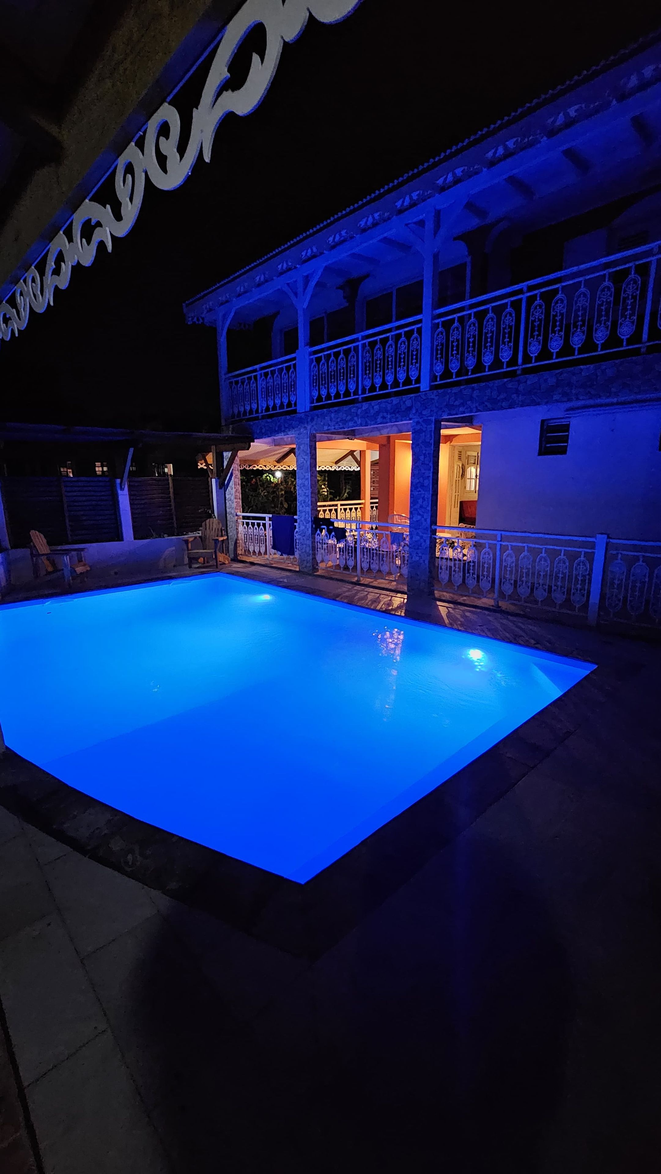 Una piscina con un área de deck iluminada en azul, un espacio de estar techado al aire libre y un edificio con balcón.