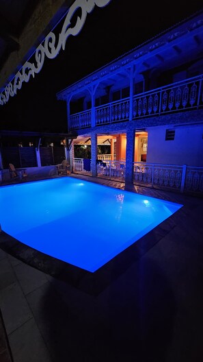 Una piscina con un área de deck iluminada en azul, un espacio de estar techado al aire libre y un edificio con balcón.