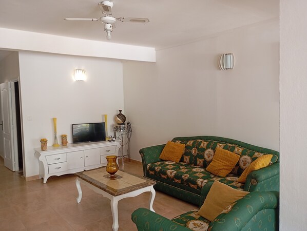 Una sala de estar con un sofá blanco, un sofá verde estampado, un mueble blanco, un televisor y un ventilador de techo.
