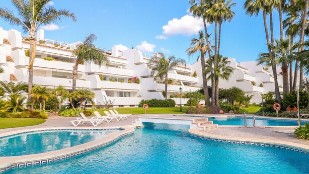 A swimming pool with lounge chairs, palm trees, and a white building with balconies. - Bahia Real apartment with large terrace (Marbella)