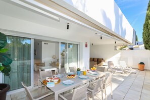 A modern outdoor dining area with a glass door leading to a living room, white furniture, and a clear view of the sky.