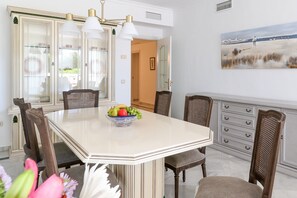 A dining area with a white table, wooden chairs, a fruit bowl, and a painting on the wall.