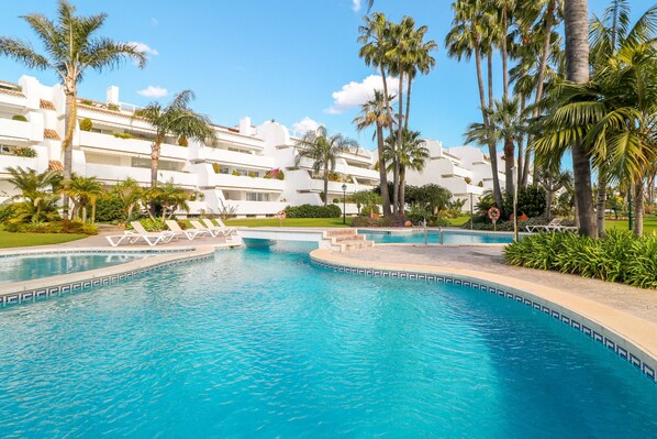 A swimming pool with lounge chairs, palm trees, and a white building with balconies.