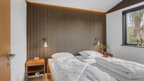 Un dormitorio con dos camas, un cabezal de pared de listones de madera, lámparas de noche, una mesita de noche y una ventana con vistas a árboles.