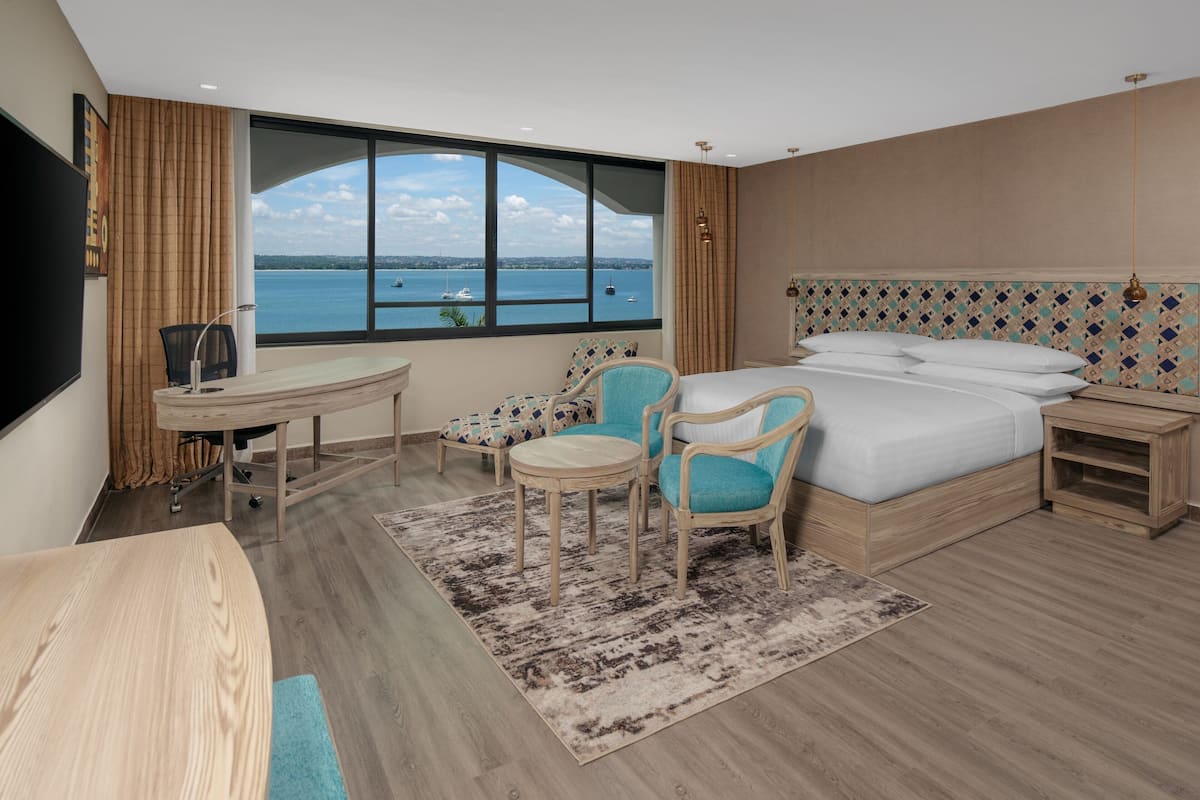 Une chambre d’hôtel dotée d’une grande fenêtre donnant sur la mer, d’un lit avec du linge de lit blanc, d’un bureau avec une chaise et d’une petite table.