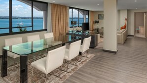 Quarto de hotel moderno com uma grande mesa de jantar, cadeiras brancas, uma mesa de vidro, vista para o mar e uma estátua de girafa.