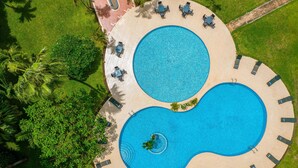 Vista aérea de um resort com duas piscinas grandes e circulares, rodeado por espreguiçadeiras e palmeiras.