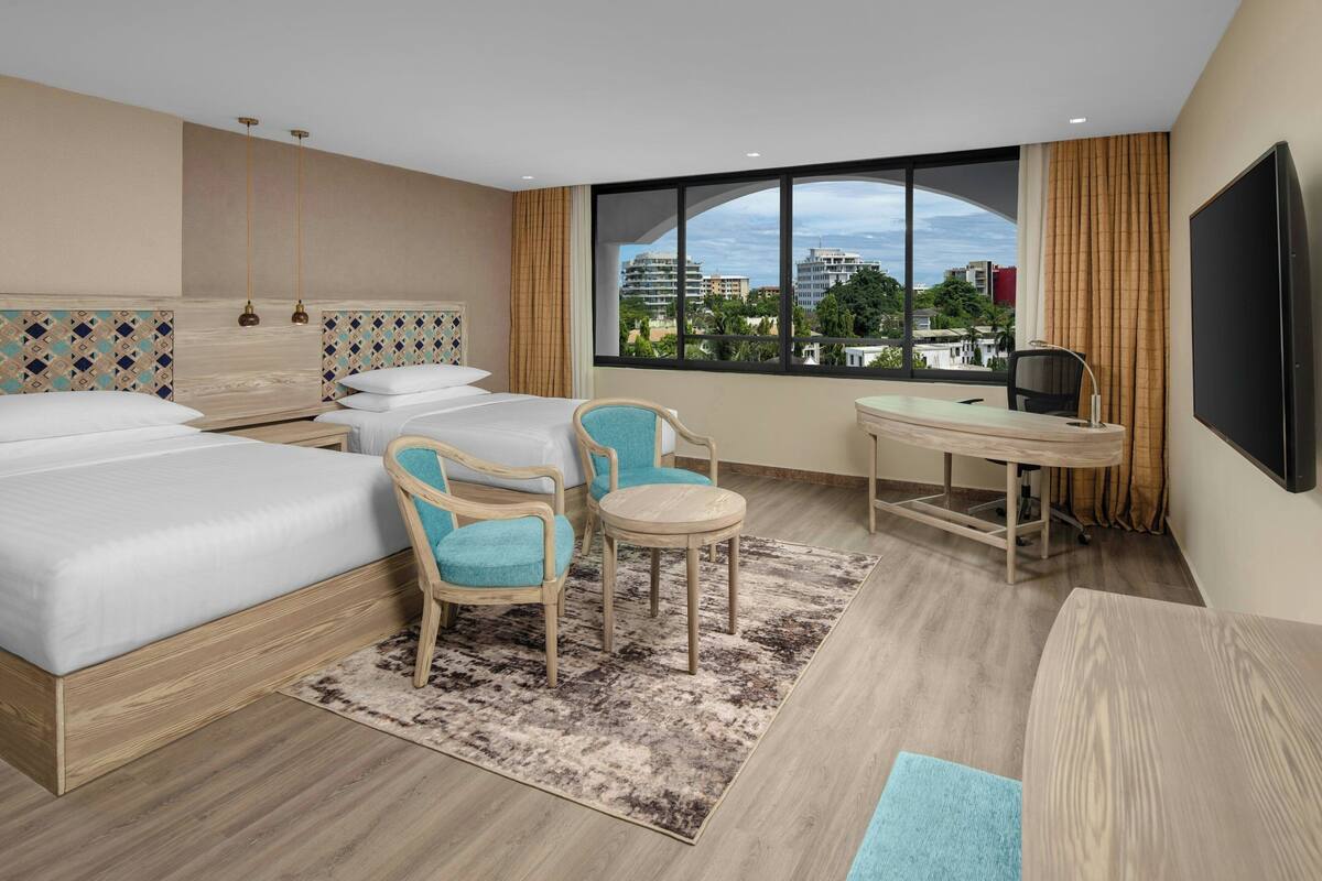 Une chambre d’hôtel avec un grand lit, deux fauteuils, une petite table, un téléviseur et une grande fenêtre donnant sur la ville.