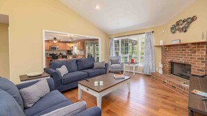 A spacious living room with a sofa, armchair, and coffee table. A fireplace and kitchen are visible in the background.