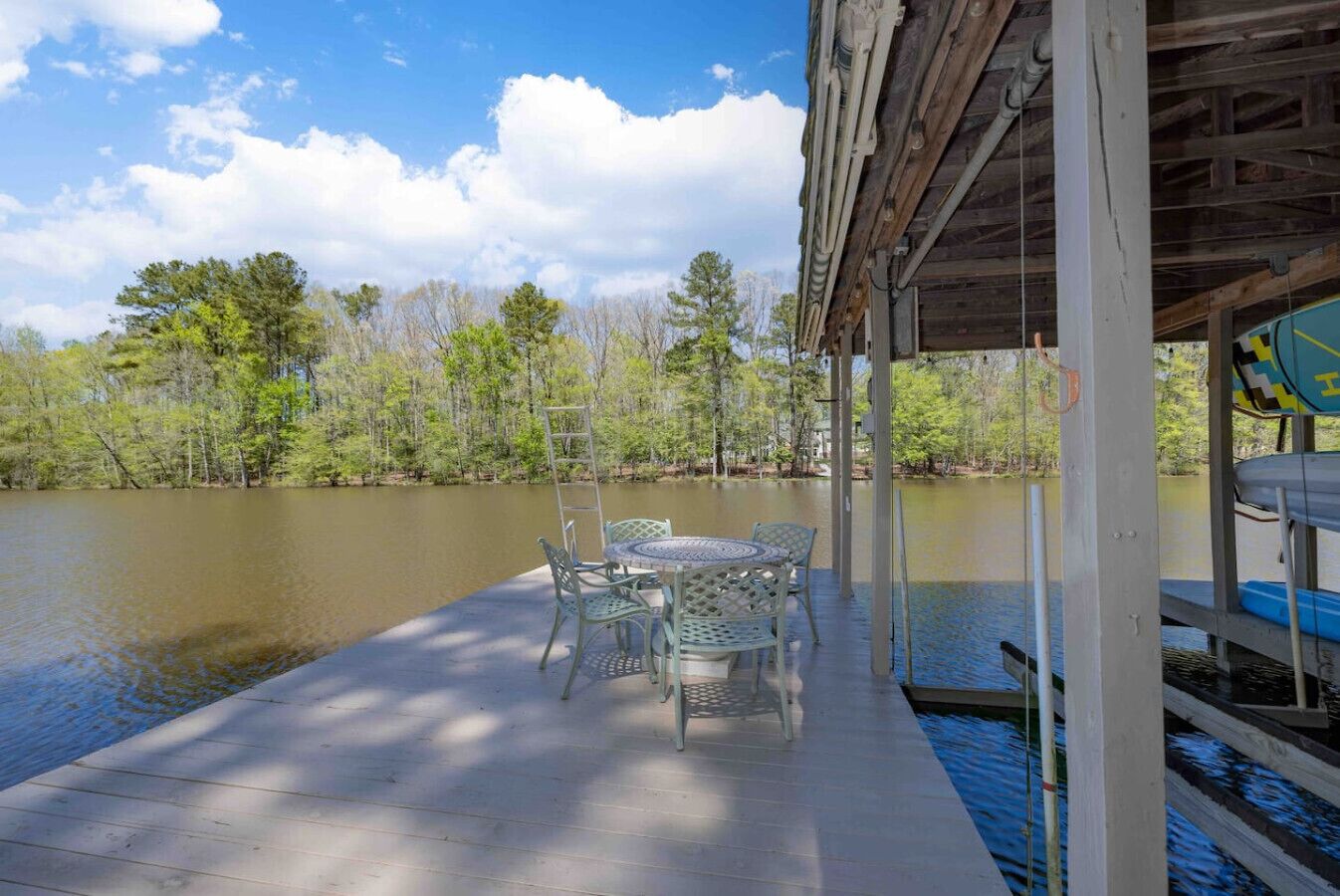 A wooden deck with a table and chairs overlooking a calm lake, surrounded by trees.