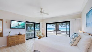 Een slaapkamer met een groot bed, een televisie, een dressoir en uitzicht op het strand door een schuifdeur.