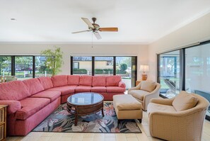 A spacious living area with a sectional sofa, a round coffee table, and two armchairs. A ceiling fan is visible, and the room has large windows offering a view of the outdoors.
