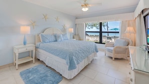 A bedroom with a bed, nightstands, a chair, a TV, and a view of the beach.