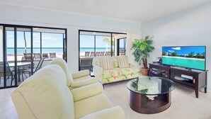 Ein geräumiges Wohnzimmer mit großem Fenster und Blick auf einen Strand, inklusive Sofa, Couchtisch und Fernseher.