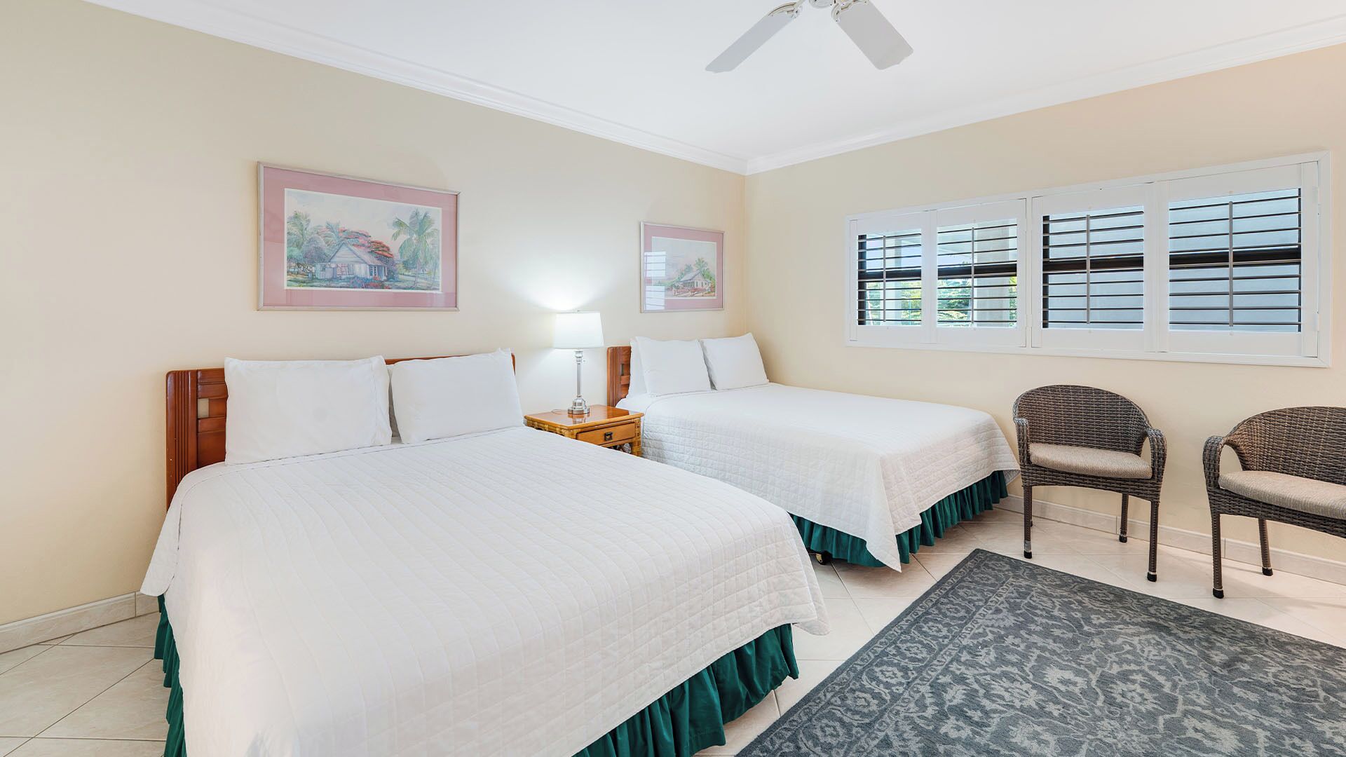 Une chambre d’hôtel avec deux lits, un ventilateur de plafond, une fenêtre avec des stores, une chaise et deux tableaux encadrés accrochés au mur.