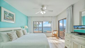 A bedroom with a bed, a television, a ceiling fan, a window with a view of the beach, and a chair. - Casa Caribe #11 - 3 Bed, 3 Bath - Oceanfront, Second Floor (Seven Mile Beach)