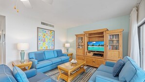 A living room with a blue sofa set, a coffee table, a television, and a bookshelf. - Casa Caribe #11 - 3 Bed, 3 Bath - Oceanfront, Second Floor (Seven Mile Beach)