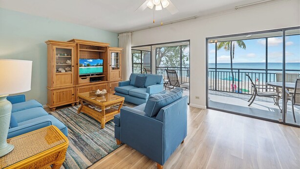 A living room with a TV, blue sofas, a wooden cabinet, and a balcony with a view of the ocean. - Casa Caribe #11 - 3 Bed, 3 Bath - Oceanfront, Second Floor (Seven Mile Beach)