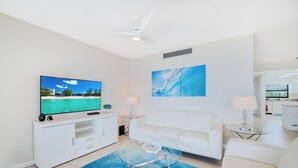 Una sala moderna con un televisor de pantalla plana, muebles blancos, una mesa de centro de vidrio y un cuadro grande en la pared.