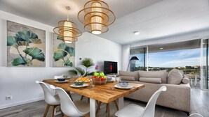 A modern living room with a wooden dining table, white chairs, a beige sofa, and large windows offering a city view. - Sonrisa | Sea view - pool - terrace (Estepona)
