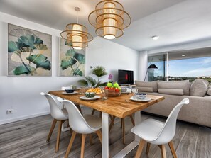 A modern living room with a wooden dining table, white chairs, a beige sofa, and large windows offering a city view. - Sonrisa | Sea view - pool - terrace (Estepona)