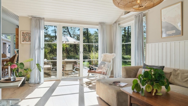 A bright, sunlit room with a large sliding glass door leading to an outdoor area, a cozy sofa, a wooden coffee table, and a wicker ceiling light. - 3 bedroom stunning home in Rødby (Rødby)