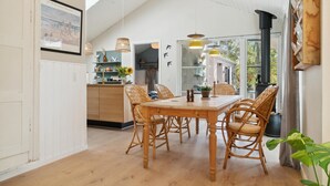 A dining area with a wooden table and wicker chairs, a kitchen with white cabinets, and a large window with a view of trees. - 3 bedroom stunning home in Rødby (Rødby)