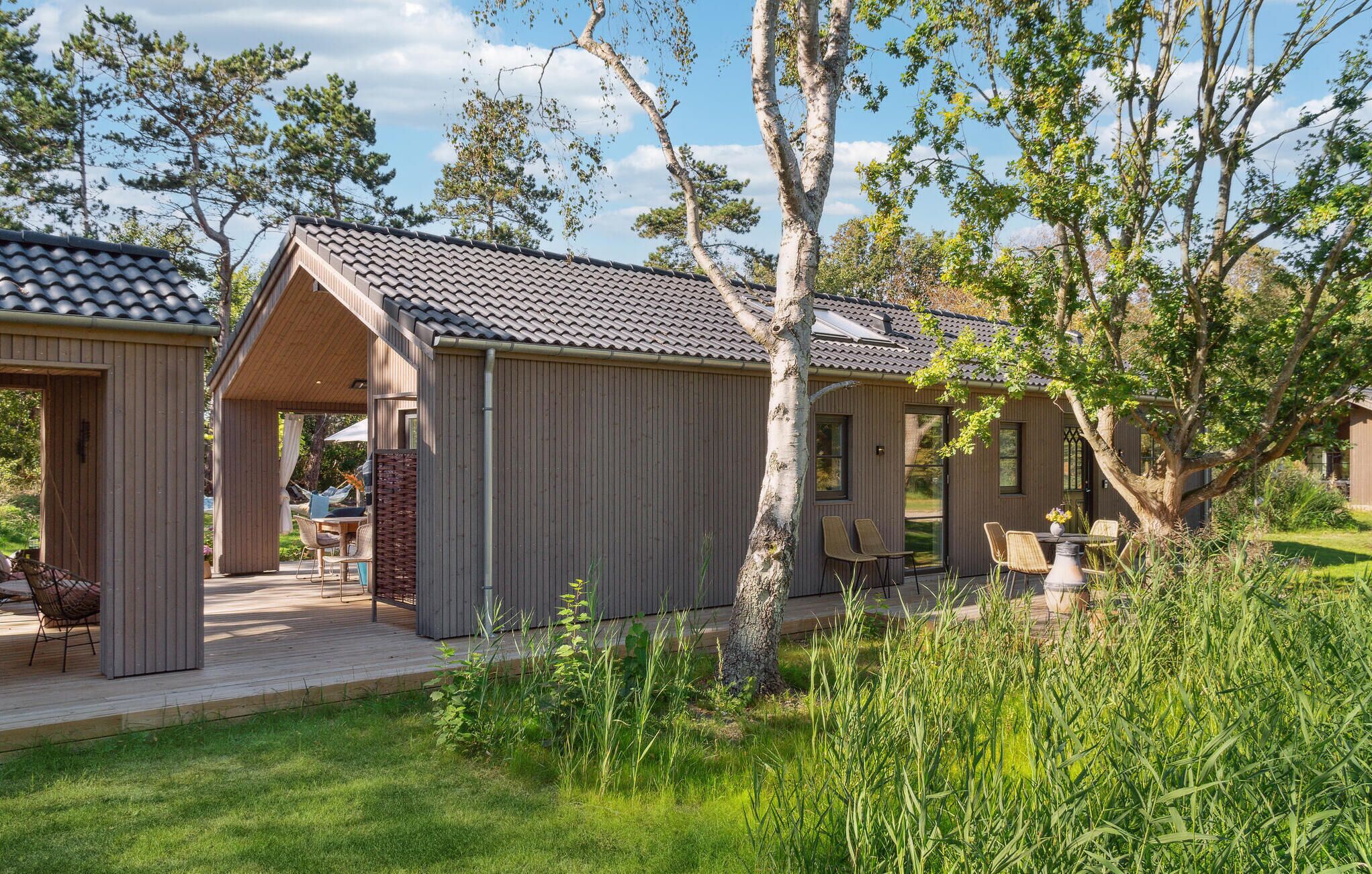 A wooden cabin with a tiled roof, a covered patio with outdoor seating, and a grassy area with trees.