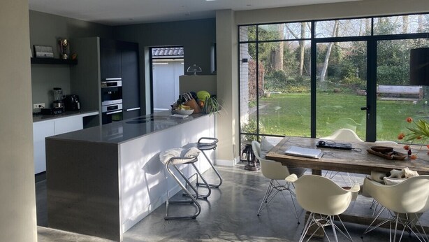 A modern kitchen with a central island, a dining area with white chairs, and large windows offering a view of a garden.