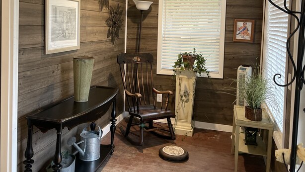 Ein gemütliches Zimmer mit Holzwänden, einem Schaukelstuhl, einem Tisch mit einer Vase und einem Fenster mit Jalousien.