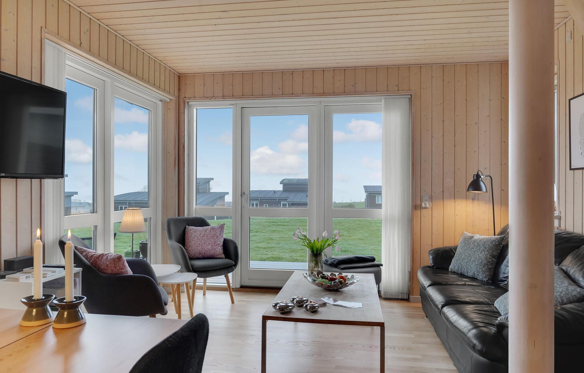 A modern living room with a large window, a flat-screen TV, a leather sofa, a coffee table, and a chair with a cushion.
