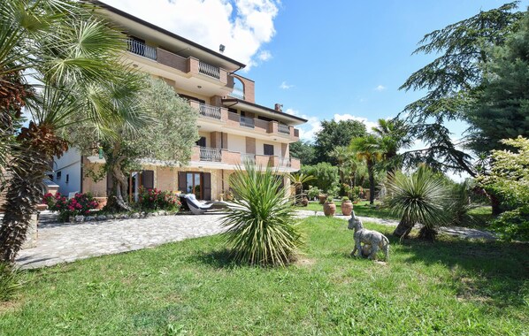 A multi-story building with balconies, surrounded by lush greenery and a stone pathway.