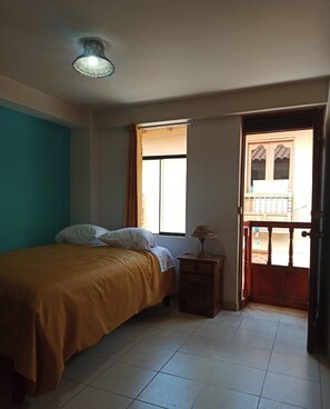A bedroom with a bed, a nightstand, a lamp, a window, and a door leading to an outdoor balcony. - Casa D Maria (Ollantaytambo)