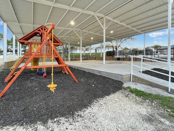 A playground with a swing set and a slide, surrounded by a covered area with a metal roof and white pillars.