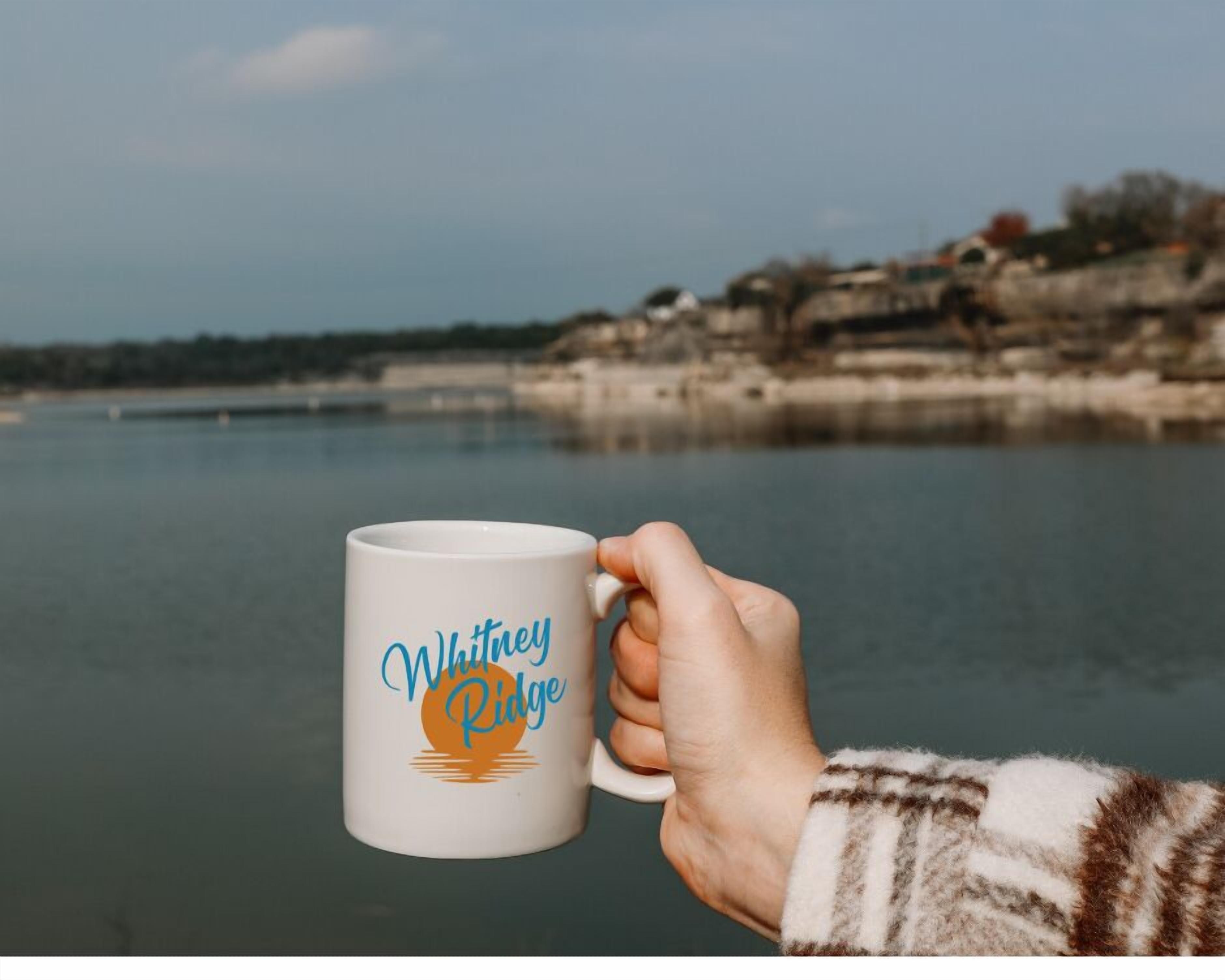 Una mano sosteniendo una taza blanca con el texto 'Whitney Ridge' y un diseño de sol.