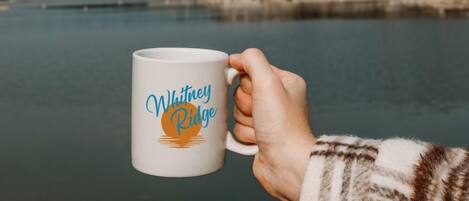 Una mano sosteniendo una taza blanca con el texto 'Whitney Ridge' y un diseño de sol.