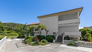 Ein zweigeschossiges Gebäude mit weißer Fassade, einem roten Ziegeldach und einem kleinen Balkon. Ein gepflasterter Weg führt zur Eingangstür, und die Umgebung ist mit verschiedenen Pflanzen und Blumen belegt.