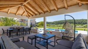 Ein überdachter Außenbereich mit Sitzgelegenheit und Blick auf einen Pool, mit Holzbalken an der Decke und einem Hängesessel.