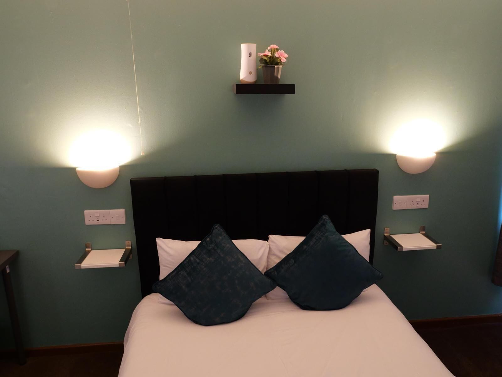 Una cama bien tendida con dos almohadas, una mesita de noche y un estante con un jarrón y flores.