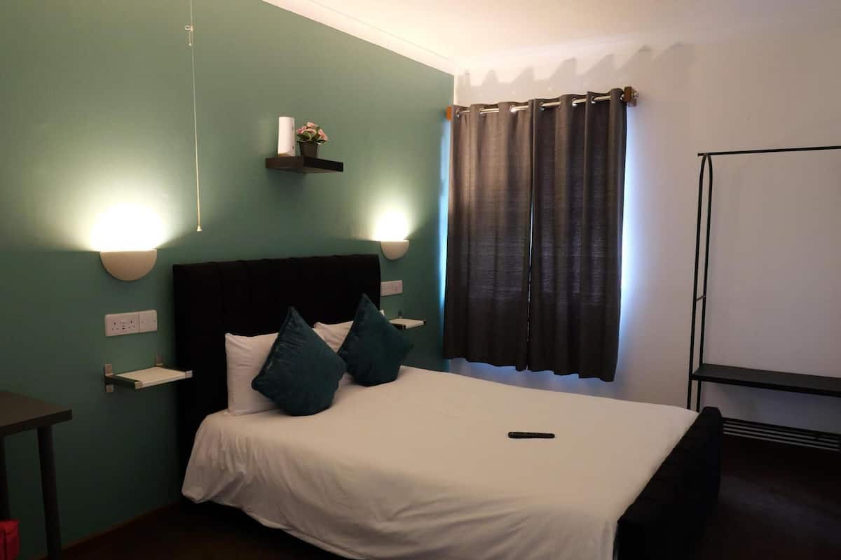 Cama bem arrumada com roupa de cama branca e duas almofadas verde-água, uma mesa de cabeceira com abajur, e uma prateleira na parede com uma pequena planta.