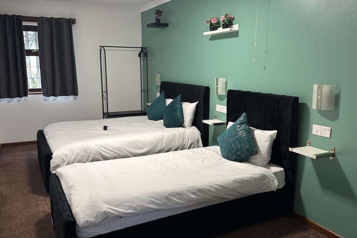 Quarto com duas camas, janela com cortinas, mesa de cabeceira e prateleira na parede com plantas.