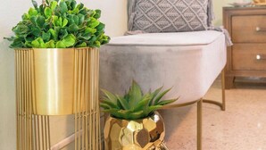 Een moderne woonkamer met een grijze bank, een decoratief kussen met een patroon en twee gouden sierpotten met planten.