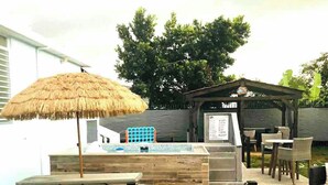 Een terras buiten met een pad van stenen, picknicktafels, een rieten parasol, een grillplaats en een overdekte zithoek.