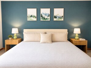 Un dormitorio ordenado con una cama grande, dos mesitas de noche con lámparas y tres cuadros con paisajes en la pared.